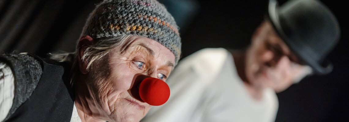 Älterer Mann mit Hut im Hintergrund. Der Schauspieler schaut den Souffleur an. Im Vordergrund ebenfalls ein älterer Mann mit Clownsnase, der Souffleur.